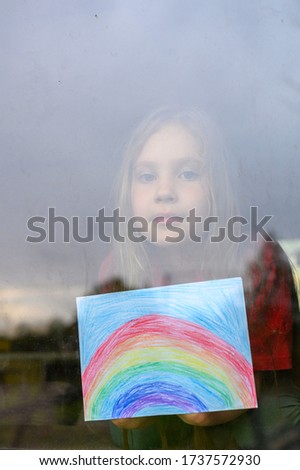 kid girl seven year old with drawing rainbow looks through the window during covid-19 quarantine. stay at home social media campaign for corona virus prevention, let's all be well. vertical image
