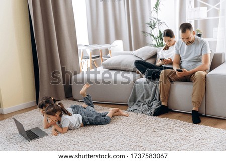 Free time at home. Two cute sisters in casual wear watching their favorite cartoon on a laptop while lying on the floor while their young parents are resting on the couch and using their smartphones