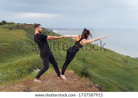 Handsome, attractive athlete in a black T-shirt and jeans holding the hand of his athletic girl. Doing acroyoga in the air outdoors. Sportswoman making step into the abyss from the cliff. Partner yoga