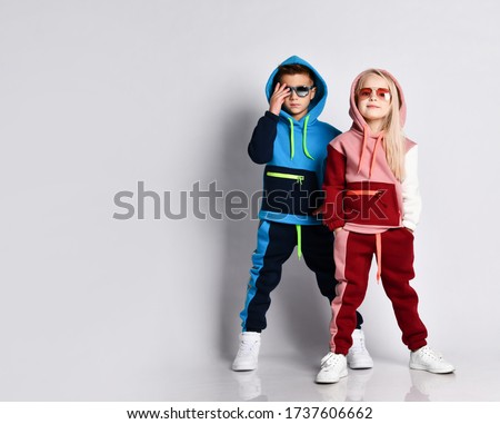 Little kids, boy and girl, in sunglasses and hoods, colorful tracksuits, sneakers. They posing isolated on white studio background. Childhood, fashion, advertising and sport. Full length, copy space Royalty-Free Stock Photo #1737606662