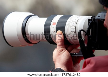 Closeup of a photographer holding onto a camera and large white telephoto lens while wearing a bright red jacket.