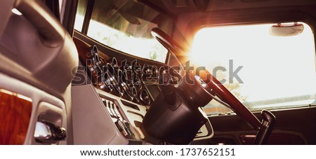 Classic american commercial semi truck with stylish chrome gauges panel and custom luxury wooden steering wheel. Luxurious and comfortable cabin of 18 wheeler truck at evening, back lit, warm filter. Royalty-Free Stock Photo #1737652151