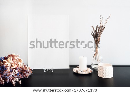 Interior design of room, blank white canvas for artist's mock-up, lips shaped vase with dry flowers bouquet, candles, white books on black table and white wall. Minimalism style.