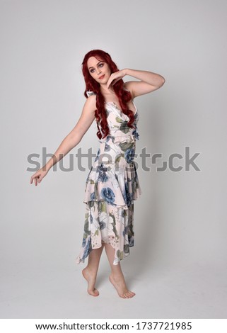 Portrait of a beautiful woman with red hair wearing  a  flowing floral gown.  full length standing pose, isolated against a studio background