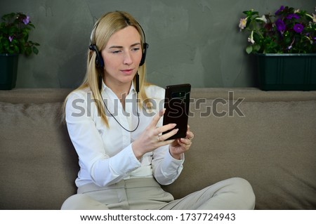 Photo of a pretty business blonde girl talking on a smartphone in headphones. Caucasian model posing at home sitting on a sofa with a smile.
