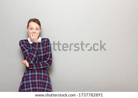 Studio portrait of shocked frightened girl with eyes widened, wearing checkered dress, biting her nails in panic, seeing something horrible or awful, standing over gray background, copy space on right