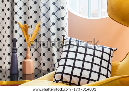 Cosy yellow armchair with striped black and white pillow by the oval window with polka dotted curtain and glass table with two vases with dry flowers.
