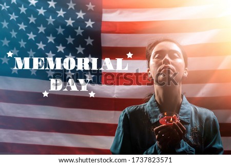 Memorial day inscription. A woman holds a candle in memory of the victims and raised her head with her eyes closed. The American flag is in the background. Concept of American holidays