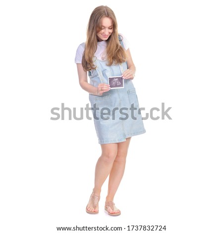 Pregnant woman in denim sundress standing looking smiling, holding ultrasound picture on white background isolation