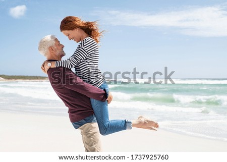 Mature couple hugging on the beach. Senior active man lifting beautiful mid woman on the beach with sea in the background and copy space. Happy retired couple hugging at seaside, looking at each other