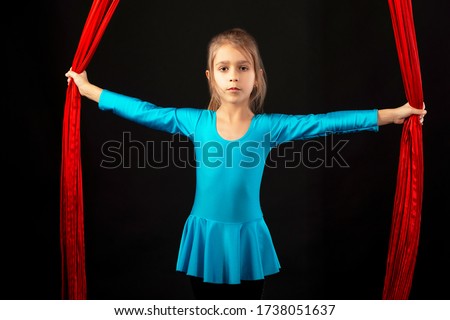 Fearless pretty little girl in a blue gymnastic suit shows a stunt aerial red ribbon on a black background. Concept of advanced acrobats gymnasts