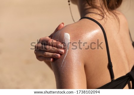 A young woman applying sun cream or sunscreen on her tanned shoulder to protect her skin from the sun. Shot on a sunny day with blurry sand in the background.  Royalty-Free Stock Photo #1738102094