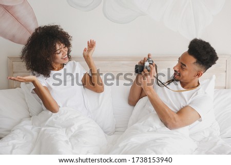 Young african loving couple girl guy in white t-shirts lying on bed doing shot on retro vintage photo camera in bedroom at home, spend time in room. Rest relax good mood quarantine lifestyle concept