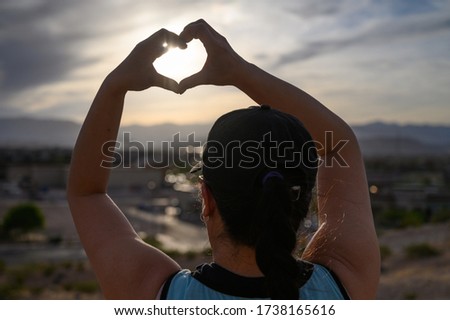 Women raising arm and hand to make a heart shape with the glowing sun in it.