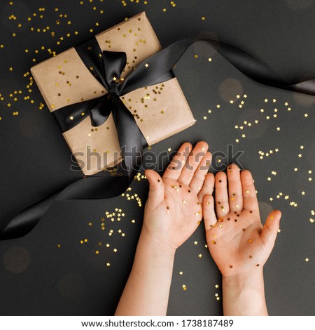 Time gifts - stylish gift box and kids hands with gold star confetti on black table. Magic stylish holiday background, black friday, sale coupon, xmas concept