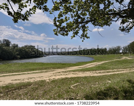 Country road. Blue river, blue sky, white clouds, green grass and leaves on a tree.