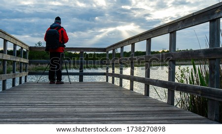 Male tourist photographer taking picture of lake. Person in red jacket on wooden pier, using tripod and camera. Lake Moszne in Poleski National Park, Poland, Europe.