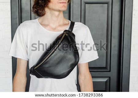 Stylish guy in a white T-shirt and a leather waist bag on a background of black doors. Designer Banana Belt. Stand straight. Convenient bag for walking and traveling. Royalty-Free Stock Photo #1738293386
