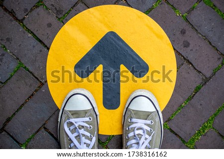 Feet wearing sneakers in front of arrow on road outdoors. Direction arrow in front of a store for the social distancing during the covid-19 pandemic. One way