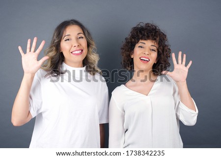Two young women standing against gray wall showing and pointing up with fingers number five while smiling confident and happy.