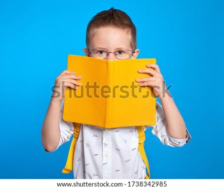 A handsome European boy with a diary and glasses looks out from behind a book. Portrait of an excellent student. Botanist. Training at school. The student laughs and smiles. Preschooler, pedagogy.