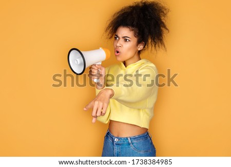 Girl shouts in megaphone and points forefinger to the side. Photo of african american girl on yellow background