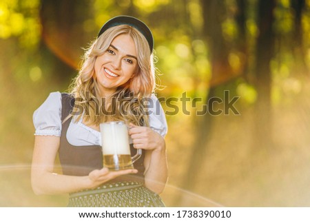 Pretty happy blonde in dirndl, traditional festival dress, holding mug of beer outdoors in the forest with blurred background. Oktoberfest, St. Patrick’s day, international beer day concept.