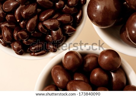 Close up detail over head view of white containers holding different sizes of dark chocolate covered nuts and fruits against a white background. Interior tempting energy sweet food.