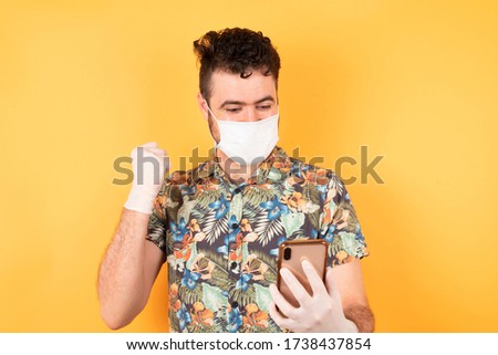 Portrait of caucasian young businessman wearing hawaiin shirt with medical mask and gloves holding in hands cell reading browsing isolated on bright vivid shine yellow background