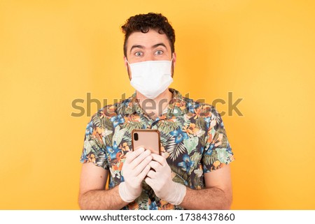Copyspace photo of cheerful nice cute charming young caucasian businessman  wearing hawaiian shirt with medical mask holding phone in her hands while isolated with bright yellow background