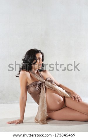 Beauty portrait of an attractive pregnant woman sitting down and and relaxing while wrapped in transparent silk fabric against a spacious plain background. Interior pregnancy beauty.