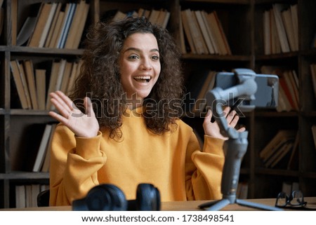 Excited hispanic teen girl social media channel blogger recording vlog on digital smartphone cam in library. School student vlogger talking looking at mobile phone on tripod shooting blog, streaming.