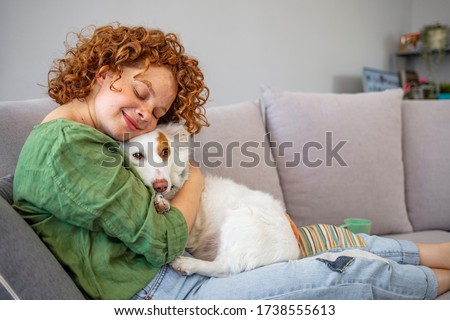 Close-up portrait of pleased girl with curly red hair embracing funny dog with eyes closed. Smiling young woman in green shirt enjoying good day and posing with pet on sofa at home. 