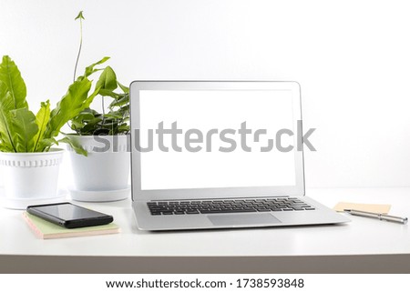 Close-up of open blank screen laptop on desk table with a smartphone, note pad, pen and potted house plant, white wall background. Modern workspace.