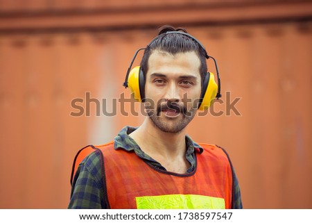 Cheerful factory worker man wearing noise reduction ear muffs and looking at camera with joy containers box background, Happiness and protection concept