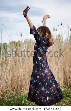Picture of young blond woman, wearing dark boho dress dancing in front of dry yellow grass in field. Hippie musician relaxing in natural environment.