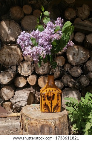 Still life, a bouquet of lilacs in a brown vintage vase, against the background of firewood in the sunshine.