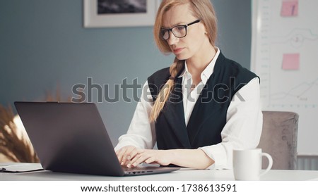 Busy mature blond businesswoman working on laptop sitting at desk with whiteboard on background