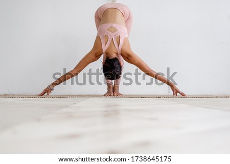 Cute slim young unidentified brunette woman doing yoga uttanasana while standing on rug on the floor in a pink barefoot suit. Young woman is training at home. Places for advertising