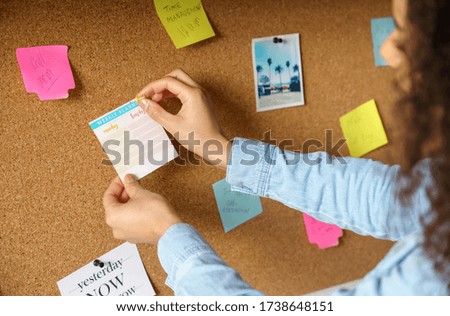 Close up over shoulder view of young african american woman pin weekly planning sticker on mood board at home office. Girl put many sticky note memo on noticeboard to organize life and work concept