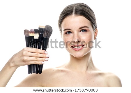 a close-up portrait of a young brunette girl with blue eyes with clear skin, with five makeup brushes in her hands, isolated on a white background. High-resolution photos