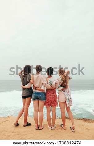 Group of four young stylish cuddiling girls looking at the ocean waves view on the beach in summer vacation. Women Friendship. Back view. Copy space