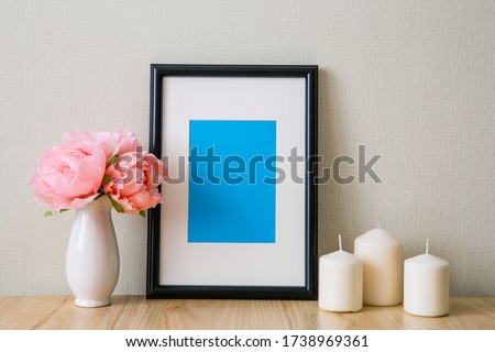 Mockup of a black vertical photo frame with Passepartout. The light interior in the Scandinavian style. Composition on the table, shelf. Pink peonies in a white vase, white candles