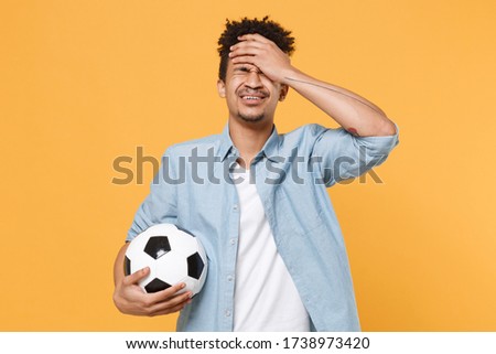 Crying african american guy football fan player in blue shirt isolated on yellow background. Sport leisure lifestyle concept. Cheer up support favorite team with soccer ball, covering face with hand