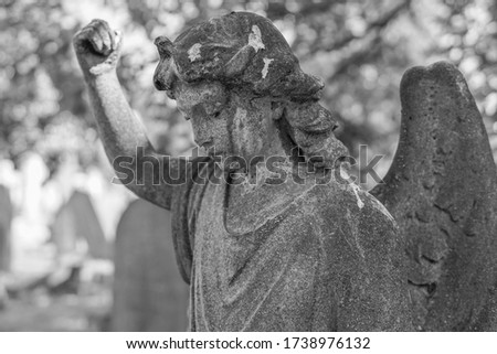 A black and white statue of an angel clenching a fist at Wandsworth Cemetery in South West London.  Image features shallow depth of field and has copy space.