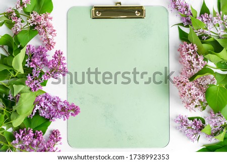Postard from frame of fresh lilac flowers and clipboard for paper on a light grey marble background. Top view.