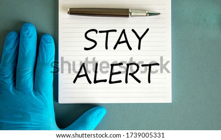 Hand in blue glove, pen, white note with words 'stay alert'.