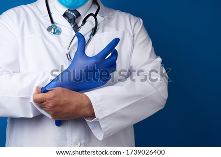 male doctor in white uniform puts on his hands blue sterile latex gloves, blue background, copy space