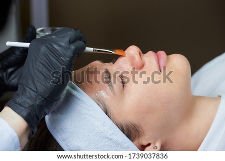 Cosmetologist wearing black disposable medical gloves uses a brush to apply a superficial transparent face peeling to a young woman. A cosmetology procedure in a beauty salon for skin cleaning. Royalty-Free Stock Photo #1739037836