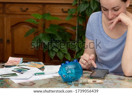 Woman calculating the bills on her smartphone. Piggy bank, credit cards, cash in different currencies and unpaid bills on the table. Concept of money saving economy for rainy days.